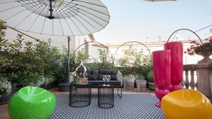 Un espacio de estar al aire libre con un sofá negro, dos mesas negras, un tapete a rayas de colores, un sillón de espuma verde, un paraguas blanco y jarrones rojos.