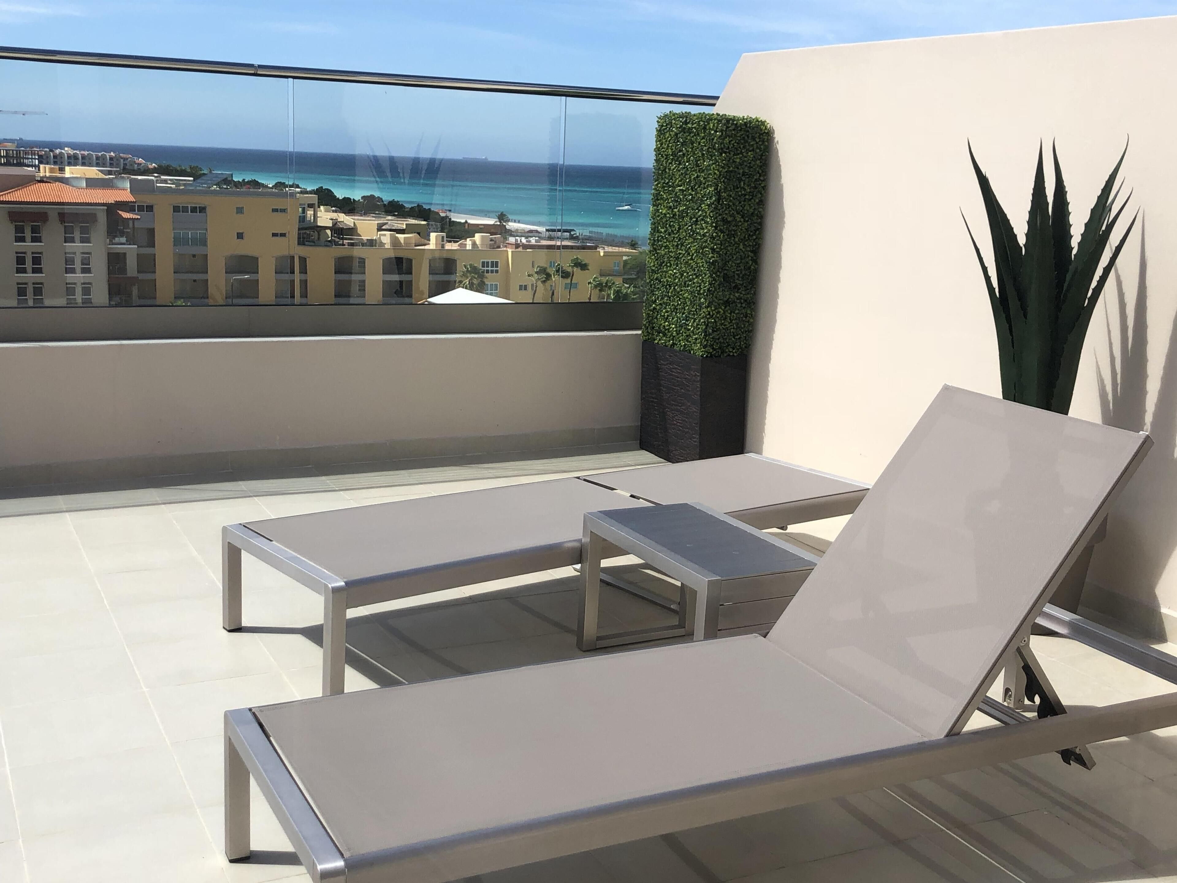 A balcony with two white lounge chairs, a potted plant, and a view of the ocean and buildings.