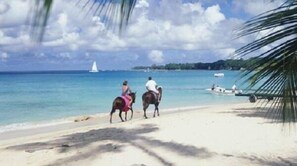 Una playa con dos personas a caballo, un velero a lo lejos y una embarcación en la orilla.