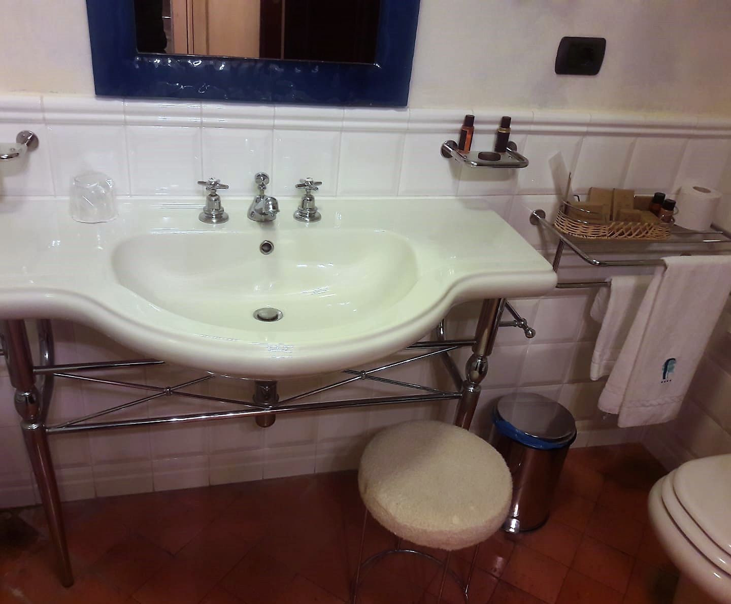 Een badkamer met een witte wastafel, chromen kraanwerk, een klein krukje, een prullenbak en een toilet.