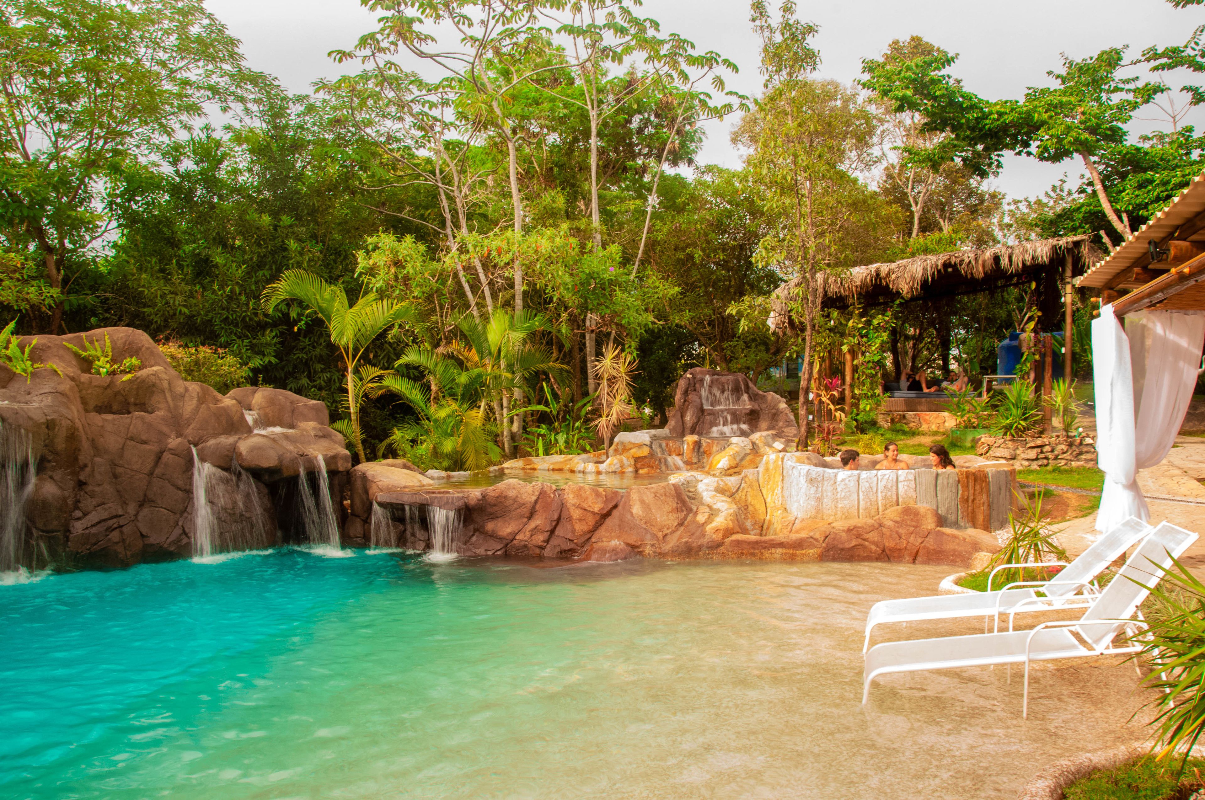 A pool area with a waterfall, lounge chairs, and a thatched-roof structure.
