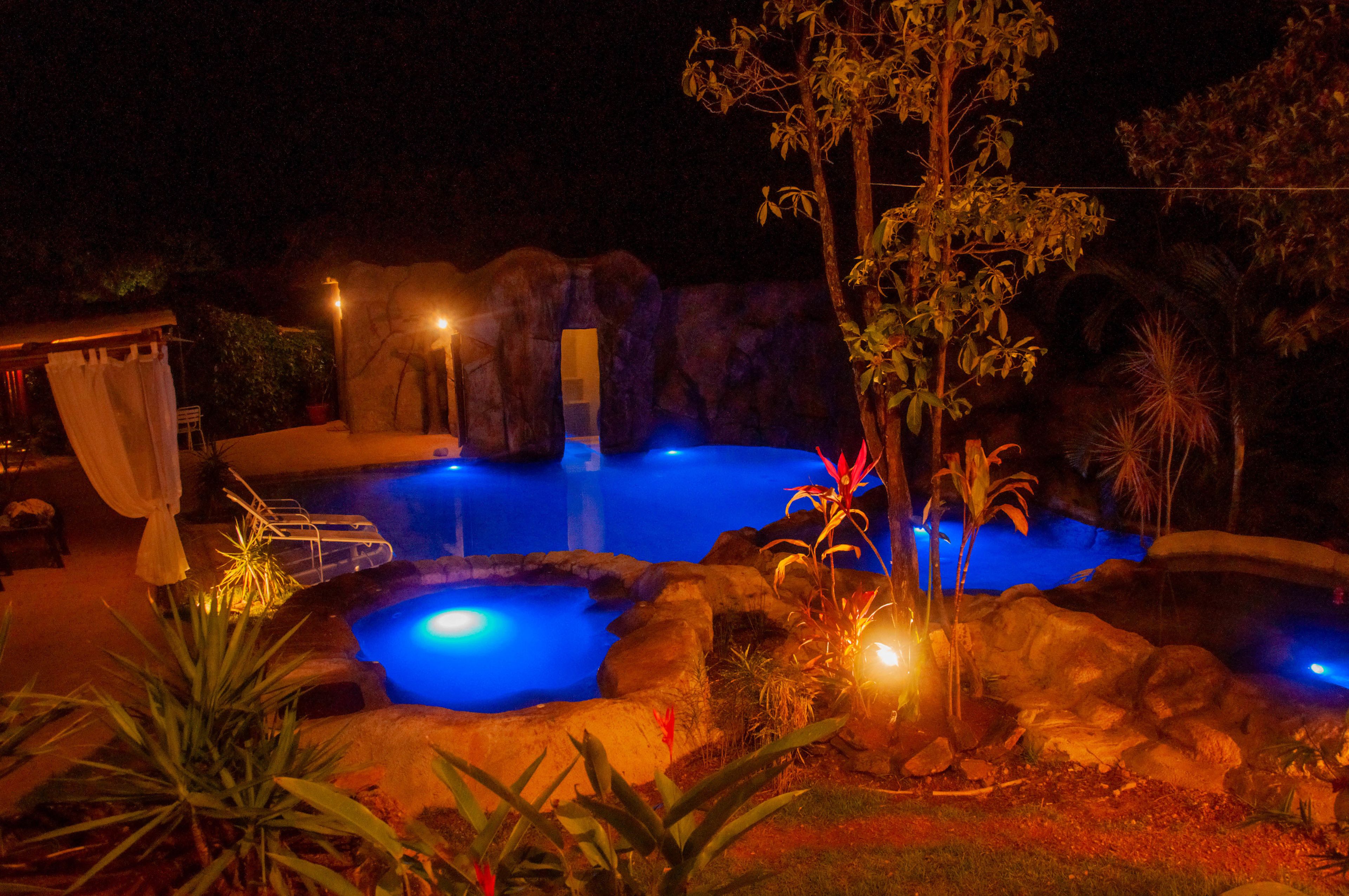 Un espace piscine nocturne avec de l’eau, des rochers et des plantes illuminés.