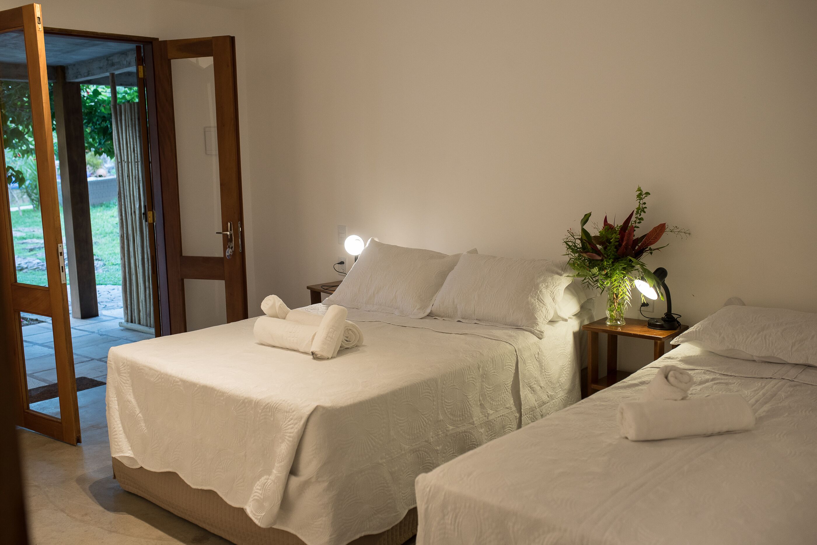 A hotel room with two beds, a nightstand with a lamp, and a view of the outdoors.