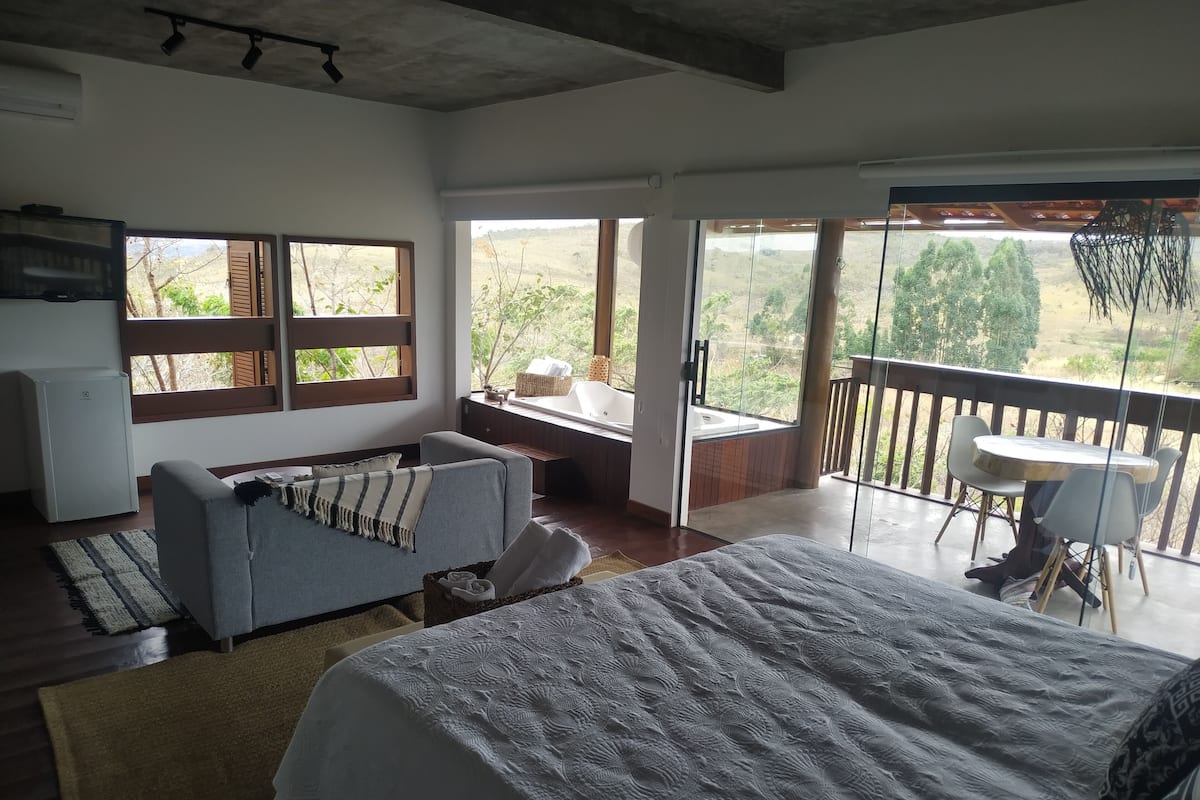 A bedroom with a large bed, a sofa, a small table, and a balcony with a view of the countryside.