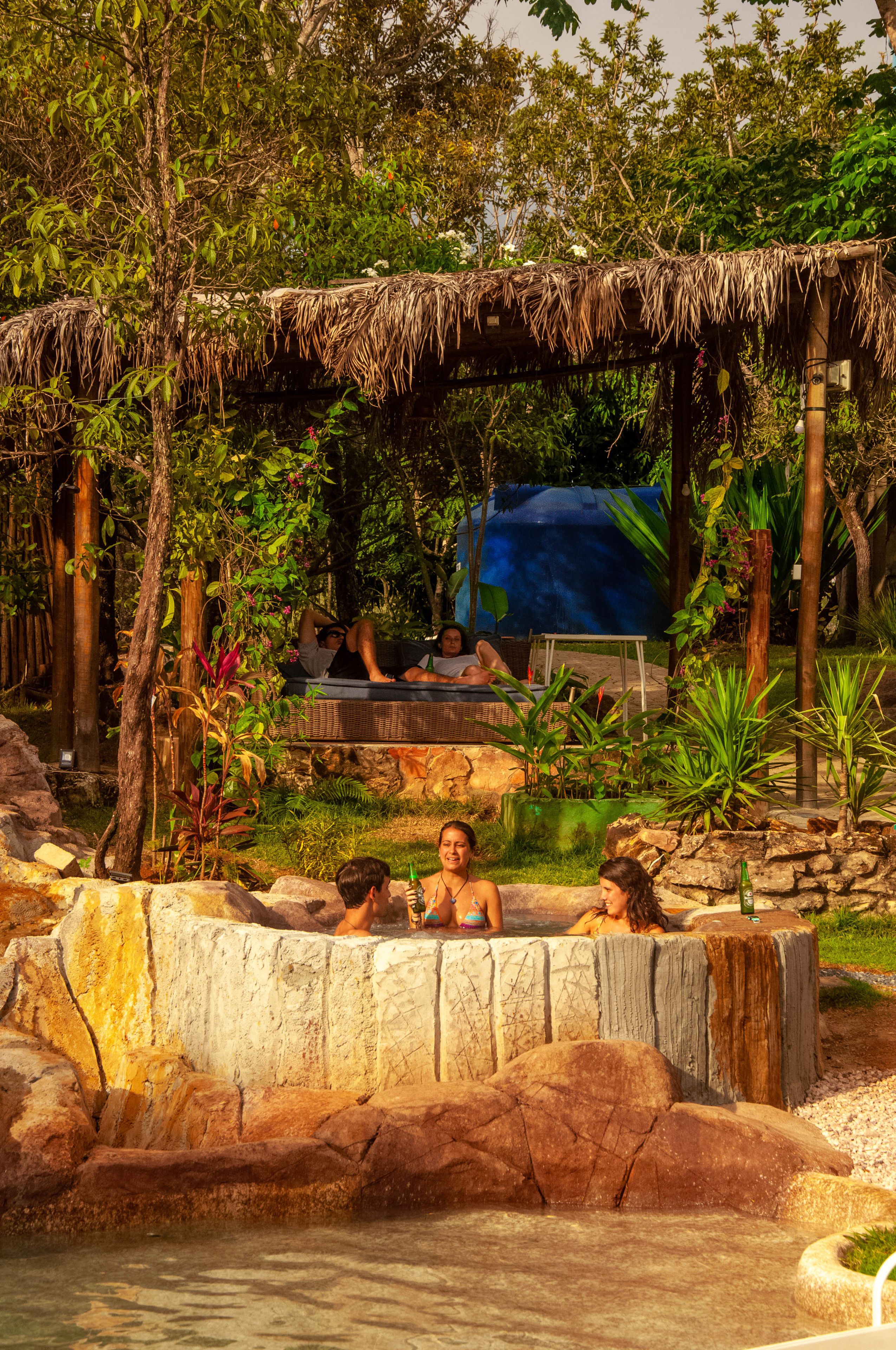 A hot spring area with a thatched roof seating area, surrounded by lush greenery and rocks.