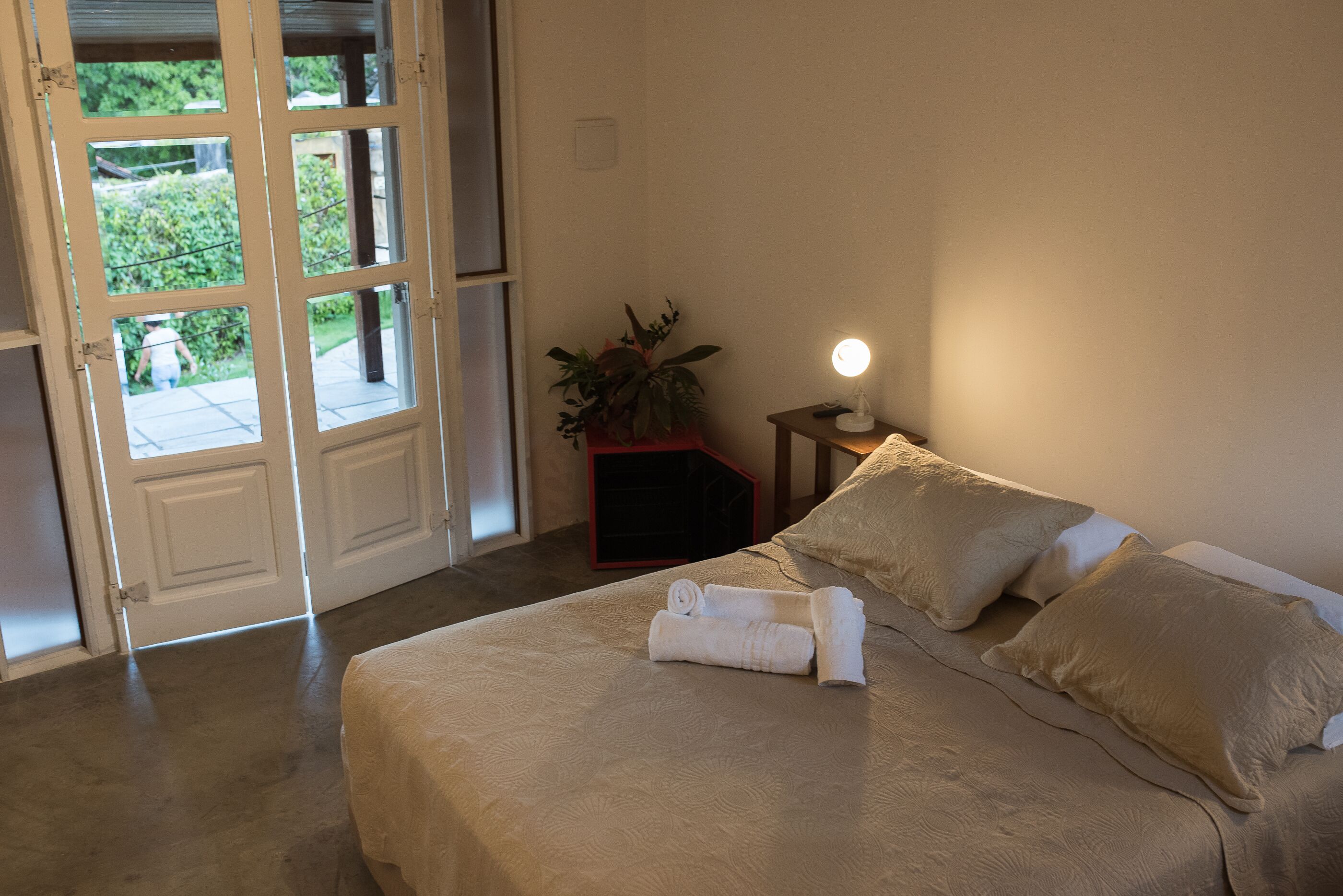 Un dormitorio con cama, mesita de noche con lámpara, planta en maceta y vistas a través de una puerta.