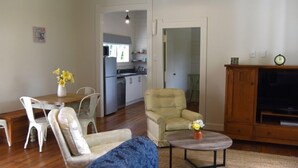 Ein Wohnzimmer mit einer Couch, einem Sessel, einem Esstisch mit Stühlen, einem TV-Schrank und einer Küche im Hintergrund.