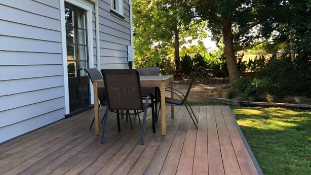 Eine Holzterrasse mit Tisch und Stühlen, umgeben von Rasen und Bäumen.