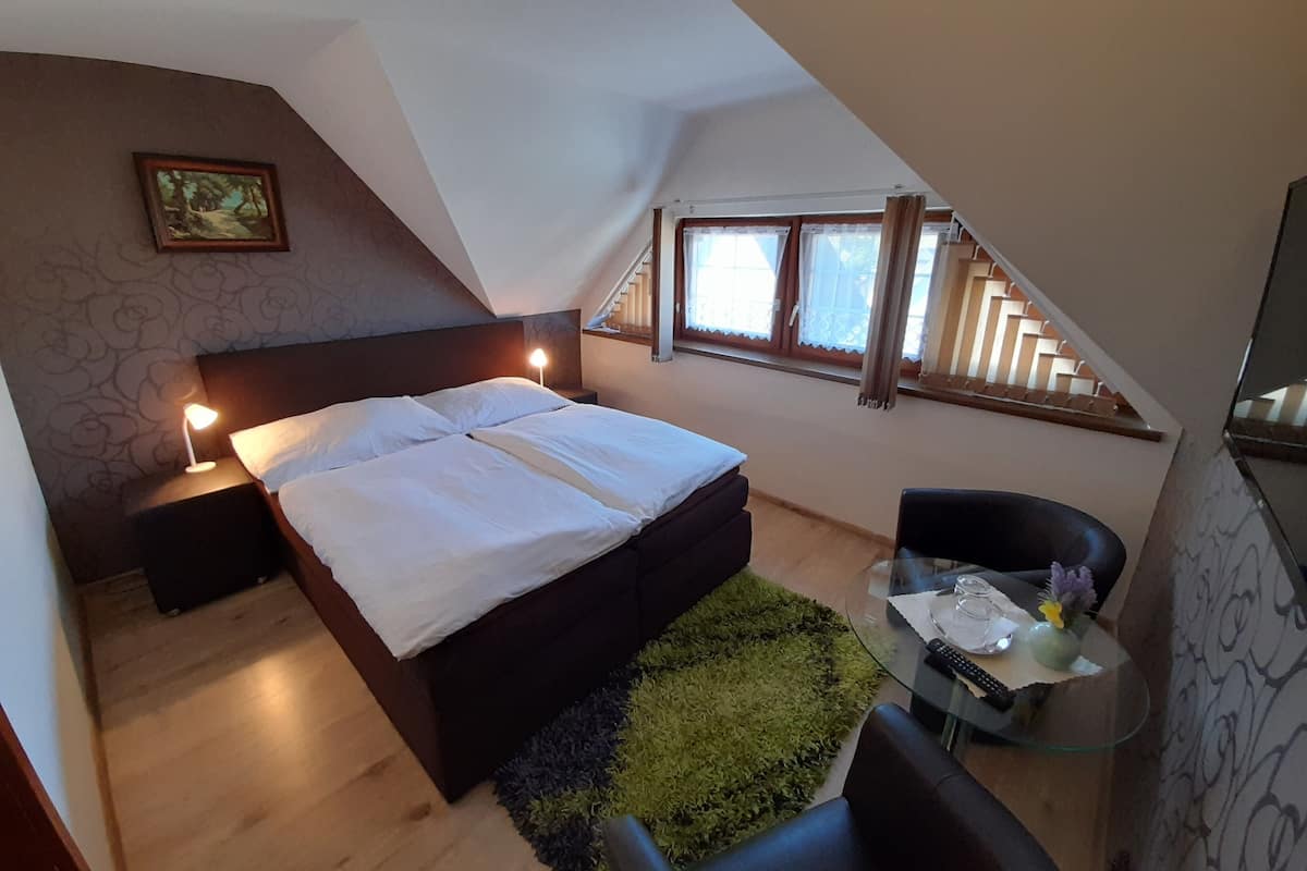 Ett sovrum med en säng, sänglampor, ett litet bord med en glasskiva, en stol och ett fönster med gardiner.