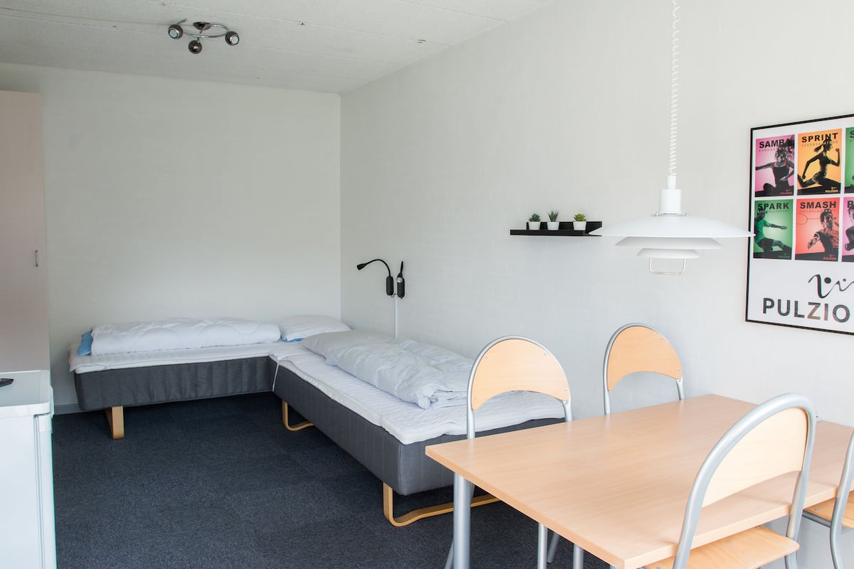 A small, neatly arranged dormitory room with two single beds, a desk, and a chair. There is a shelf with small plants and a poster on the wall.