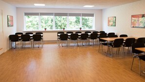 Un salón de conferencias con una larga mesa y sillas, amplias ventanas con vista a árboles y paredes de ladrillo blanco.