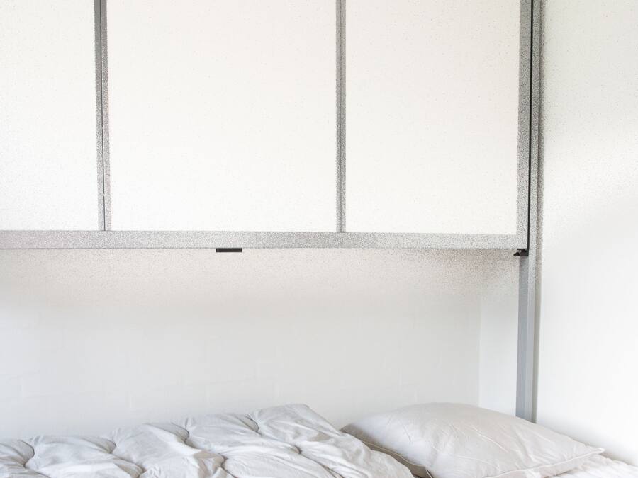 A single bed with a grey frame and white bedding, a pull-out desk, and a window with blinds.