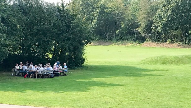 Un grupo de personas sentadas en una mesa en un campo cubierto de hierba, rodeadas de árboles.