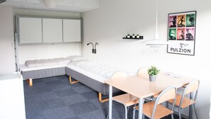 Habitación pequeña con cama, escritorio y sillas. Se aprecia un estante con plantas y un cuadro en la pared.