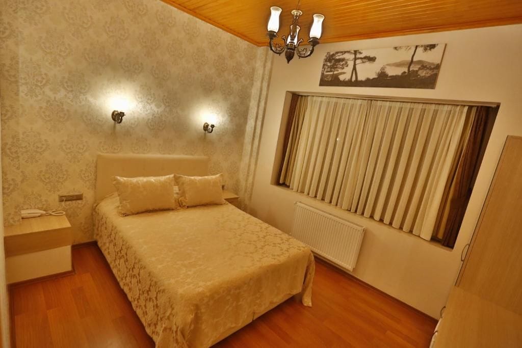 A bedroom with a bed, a nightstand, a wall-mounted light fixture, a radiator, and a window with curtains.