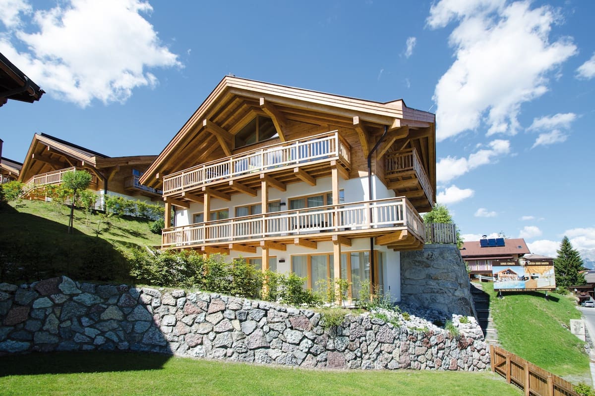 A large wooden chalet with multiple balconies, stone walls, and a hillside setting.