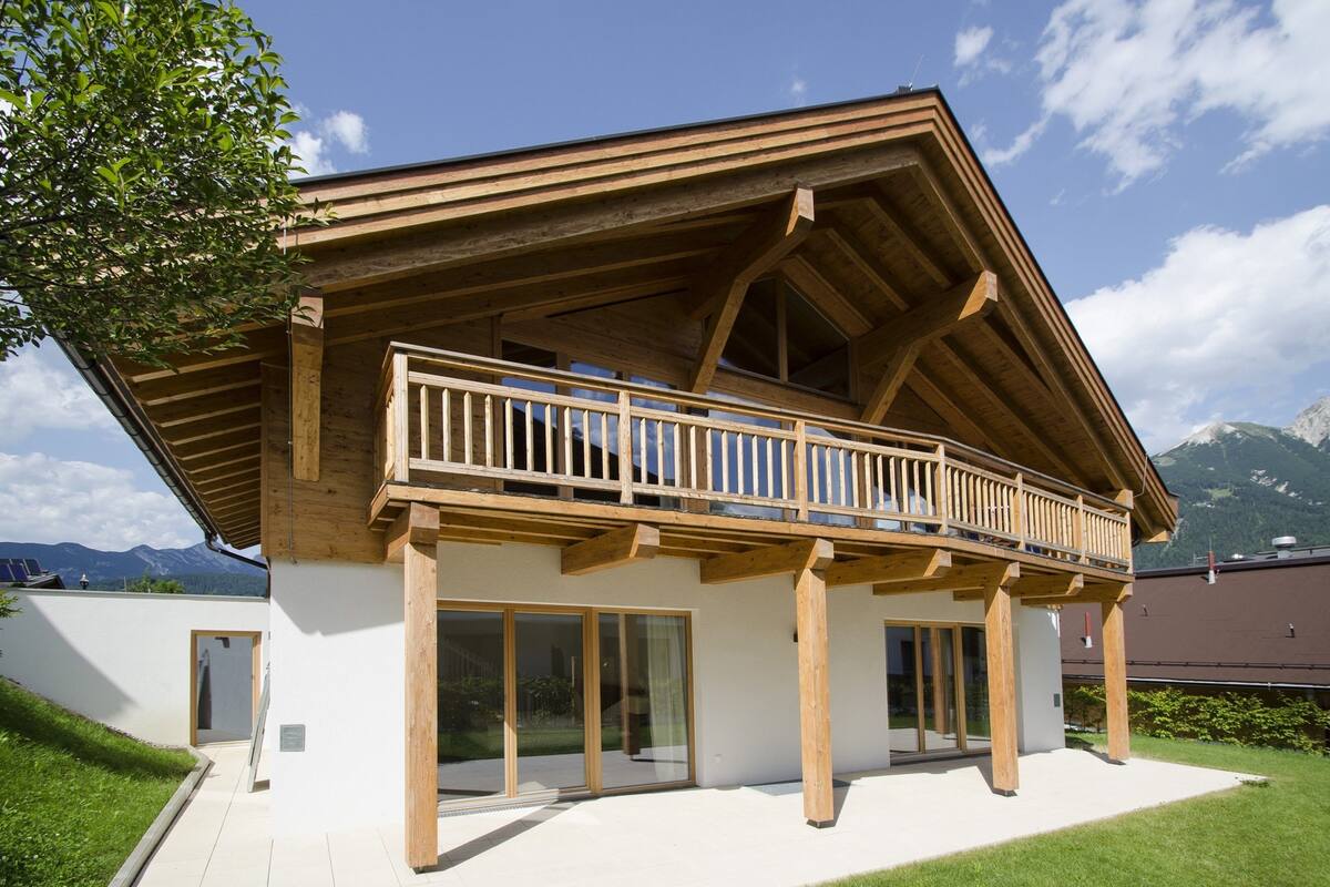 A modern wooden building with a balcony, large glass doors, and a mountain backdrop.