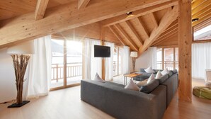 A spacious living area with a wooden ceiling, a grey sofa, a flat-screen TV, and large windows. - Mountains Chalets (Seefeld in Tirol)