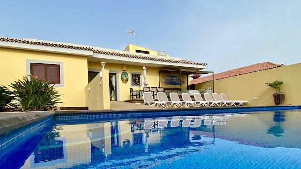 A swimming pool with clear blue water, surrounded by lounge chairs and a building with a tiled roof. - House / Villa - LAS GALLETAS (LAS GALLETAS)