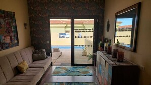 A living room with a sofa, a coffee table, a patterned rug, a cabinet with decorative items, and a view of a building through a glass door. - House / Villa - LAS GALLETAS (LAS GALLETAS)