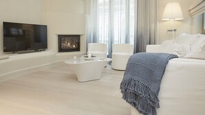 Una sala de estar moderna con un televisor de pantalla plana, chimenea, muebles blancos y un ventanal con cortinas translúcidas.