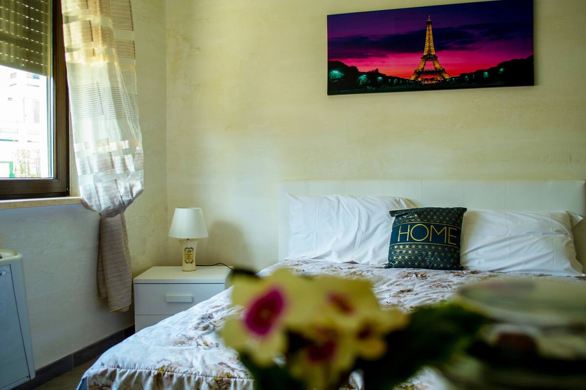 A bedroom with a bed, a nightstand, a lamp, a window with curtains, and a painting of the Eiffel Tower.
