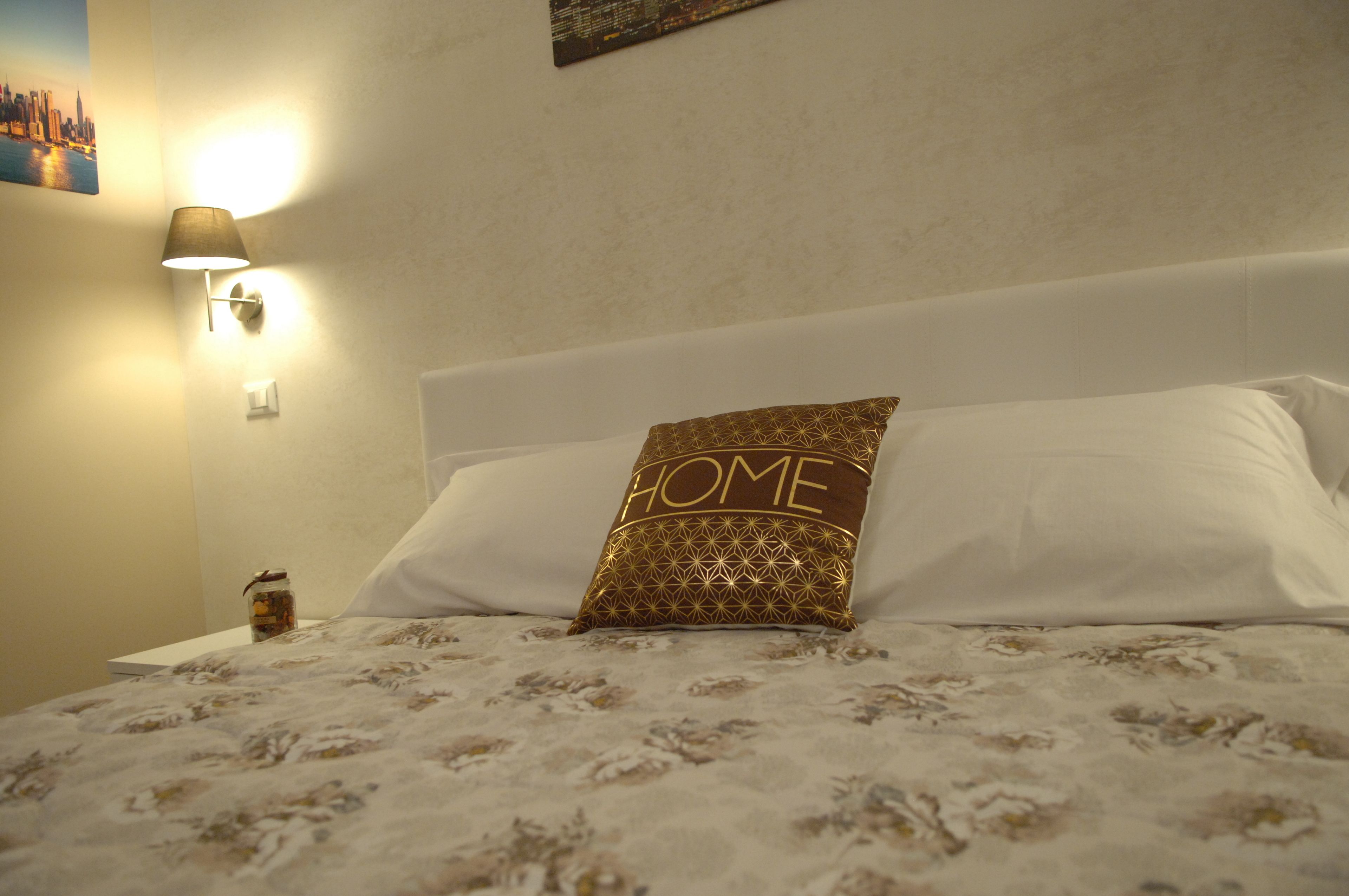 Un lit bien fait, recouvert d’une couvre-lit à motifs, un oreiller sur lequel est écrit le mot « MAISON », et une lampe fixée au mur.