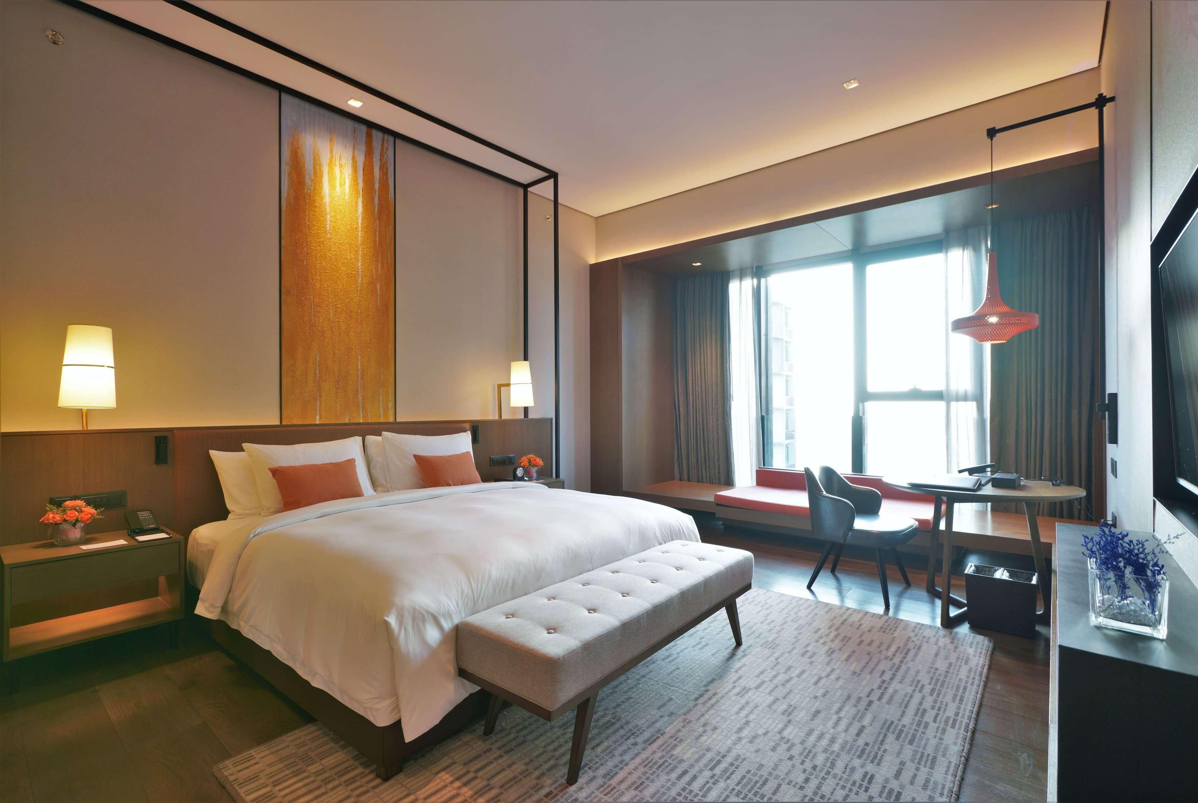 A modern hotel room with a large bed, bedside lamps, a bench, a desk, and a large window with curtains.