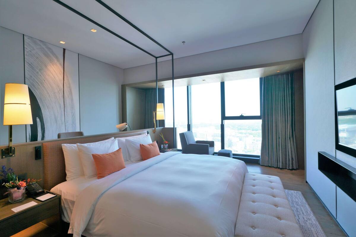 A modern hotel room with a large bed, a desk with a lamp, a chair, and a large window offering a city view.