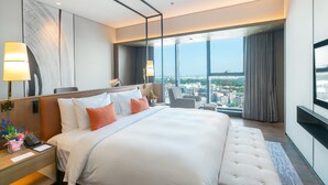 Habitación de hotel moderna con una cama grande, un escritorio con lámpara, una silla y vistas a la ciudad a través de amplios ventanales.