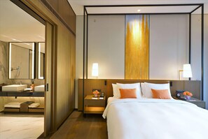 Una habitación de hotel con una cama grande, una mesita de noche con lámpara, un tocador con lámpara, un baño con lavamanos y espejo, y un panel de pared de madera.