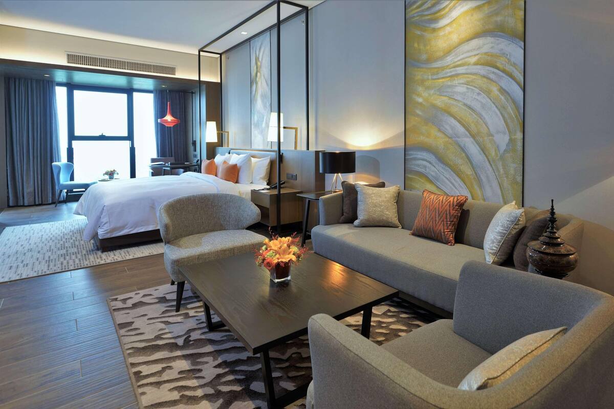 A modern hotel room with a bed, sofa, armchairs, a coffee table with flowers, a desk, and a large abstract painting on the wall.