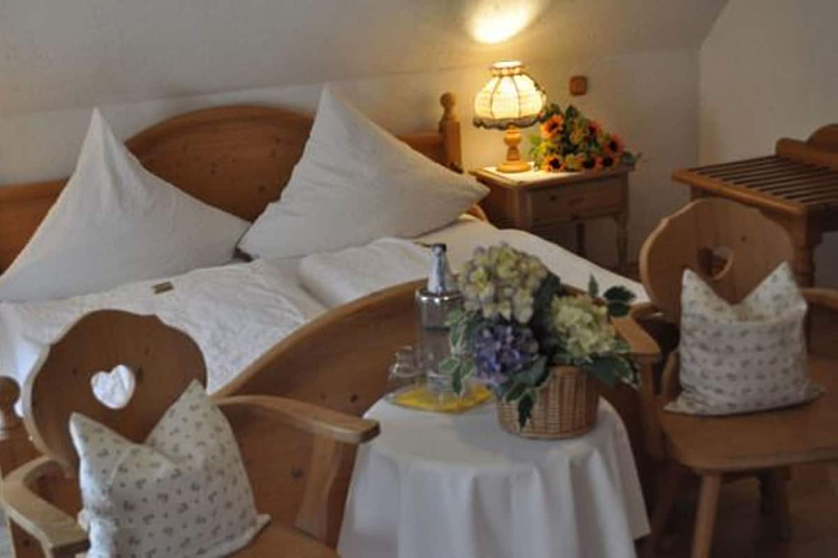 A neatly arranged bedroom with a bed, two chairs, a bedside table with a lamp, and a small table with a vase of flowers.