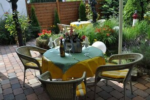 A patio with a table set for two, surrounded by wicker chairs, and a small garden with various plants and a decorative fence in the background. - Bei Hölzchen (Hannover)