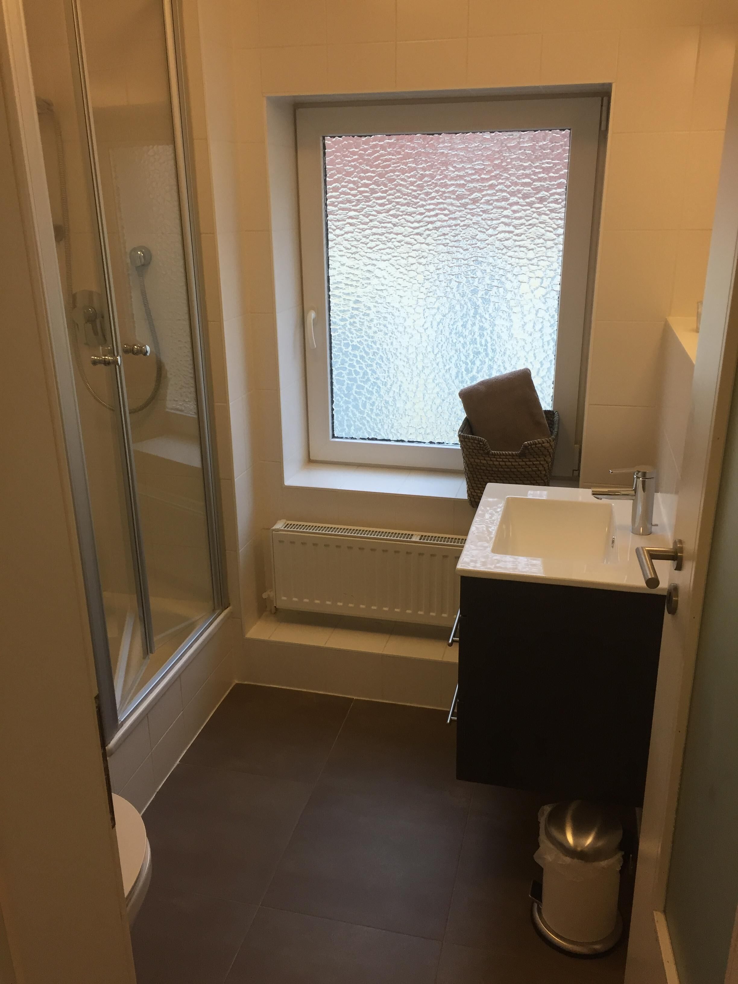 Un bany petit amb plat de dutxa de vidre, una finestra amb estoreta texturada, un radiador, un lavabo i un vàter.