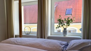 A bedroom with a bed, a window with curtains, and a view of a building with a red roof. - Cozy and charmingly furnished 30sqm apartments in a central location in Husum (Husum)