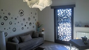 Uma sala de estar moderna com um sofá cinzento, uma parede decorativa com padrões circulares, uma luminária pendente e uma janela com um painel geométrico.