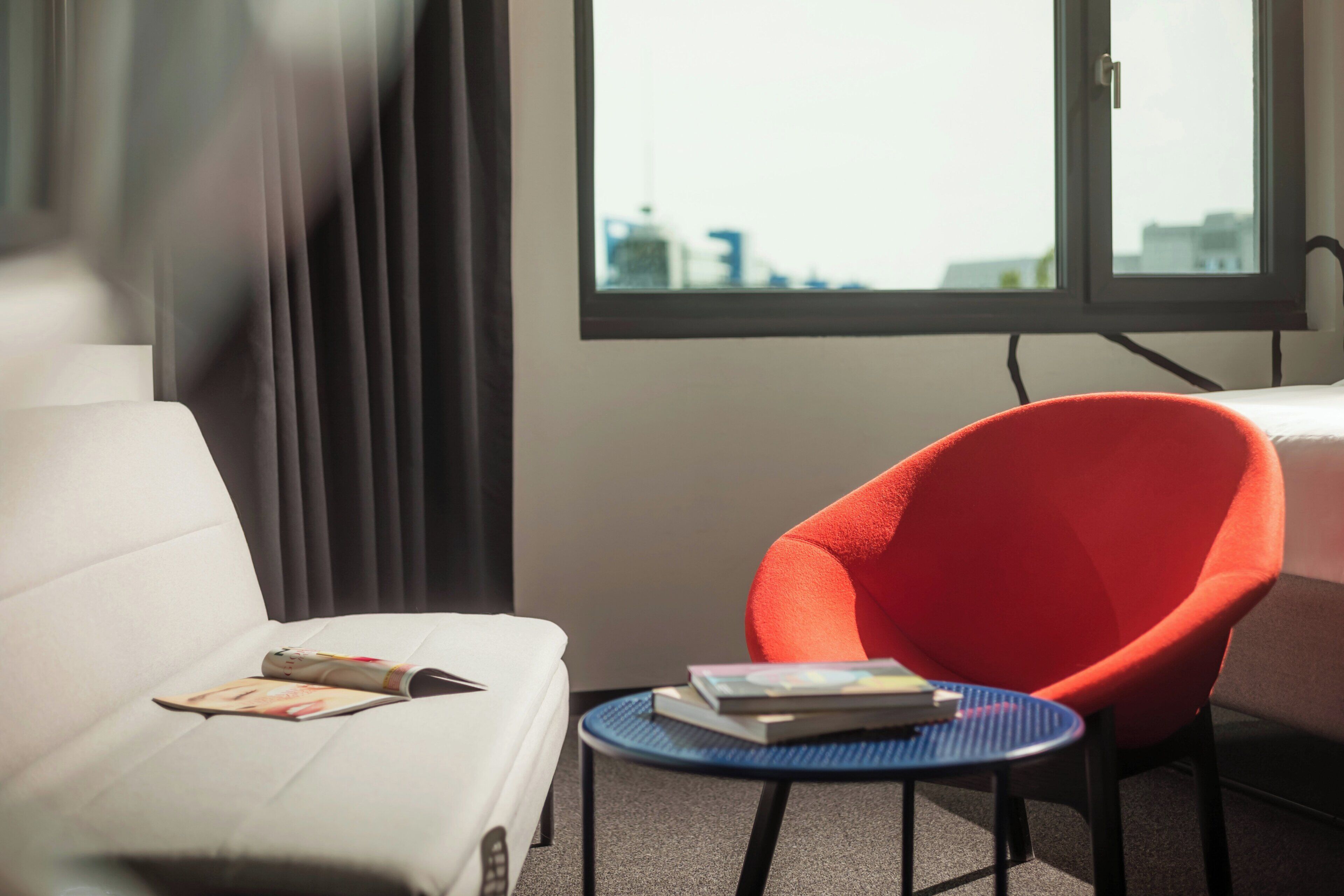 Ein Hotelzimmer mit einem weißen Sofa, einem roten Sessel, einem kleinen runden Tisch mit Büchern und einem Fenster mit Blick auf die Stadt.