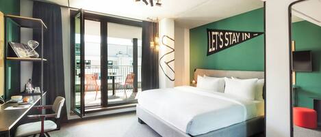 Habitación de hotel con cama, escritorio, silla, puerta corrediza de vidrio a un balcón y un letrero motivacional en la pared.