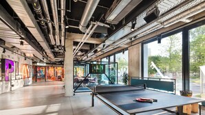 Un espacio interior con mesa de ping pong, tuberías del techo visibles y una zona de gimnasio al fondo.