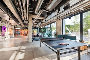 Un espacio interior con mesa de ping pong, tuberías del techo visibles y una zona de gimnasio al fondo.