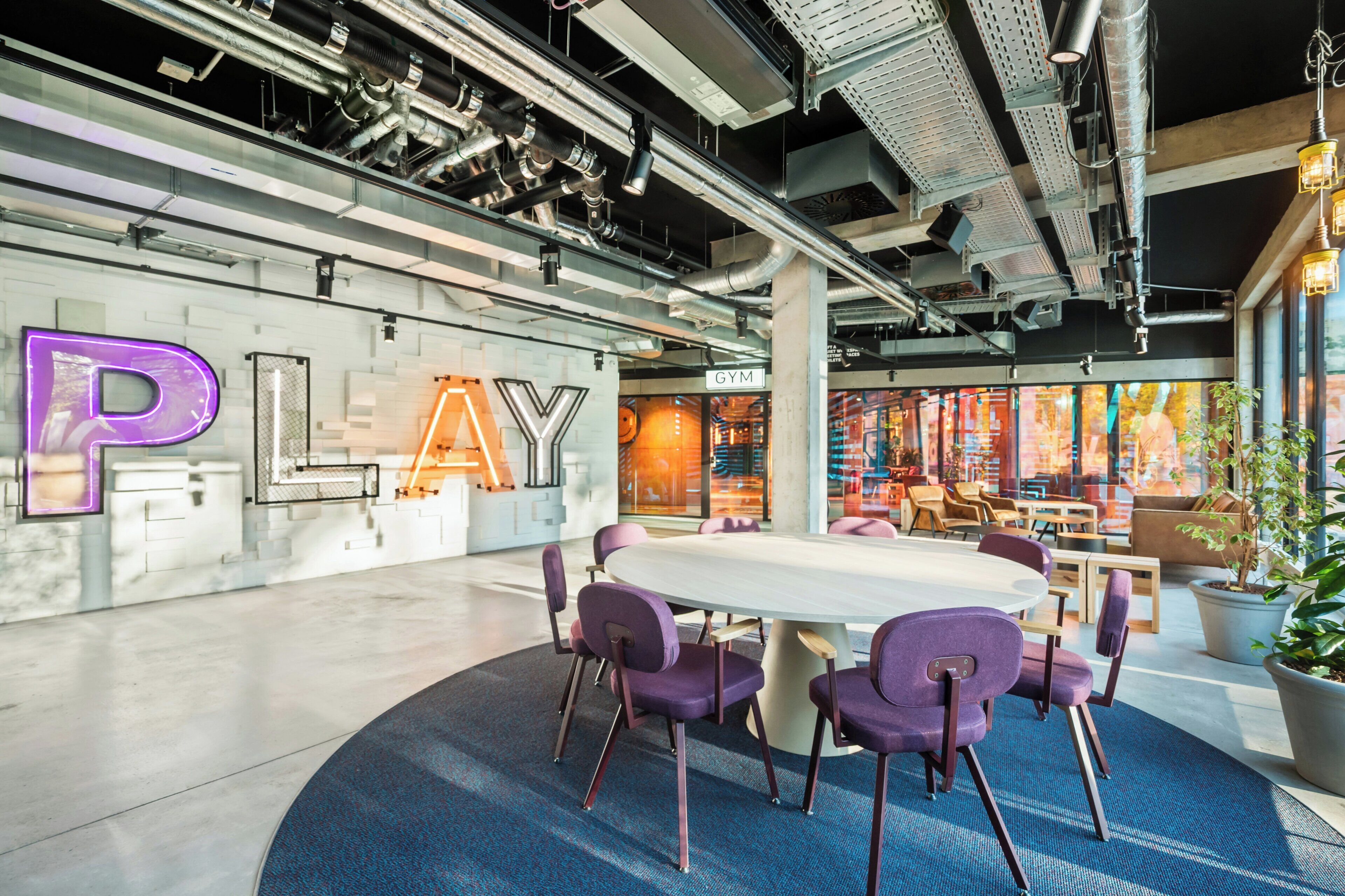 Un espacio interior moderno con una mesa grande rodeada de sillas moradas, un gimnasio visible y elementos de techo de estilo industrial.