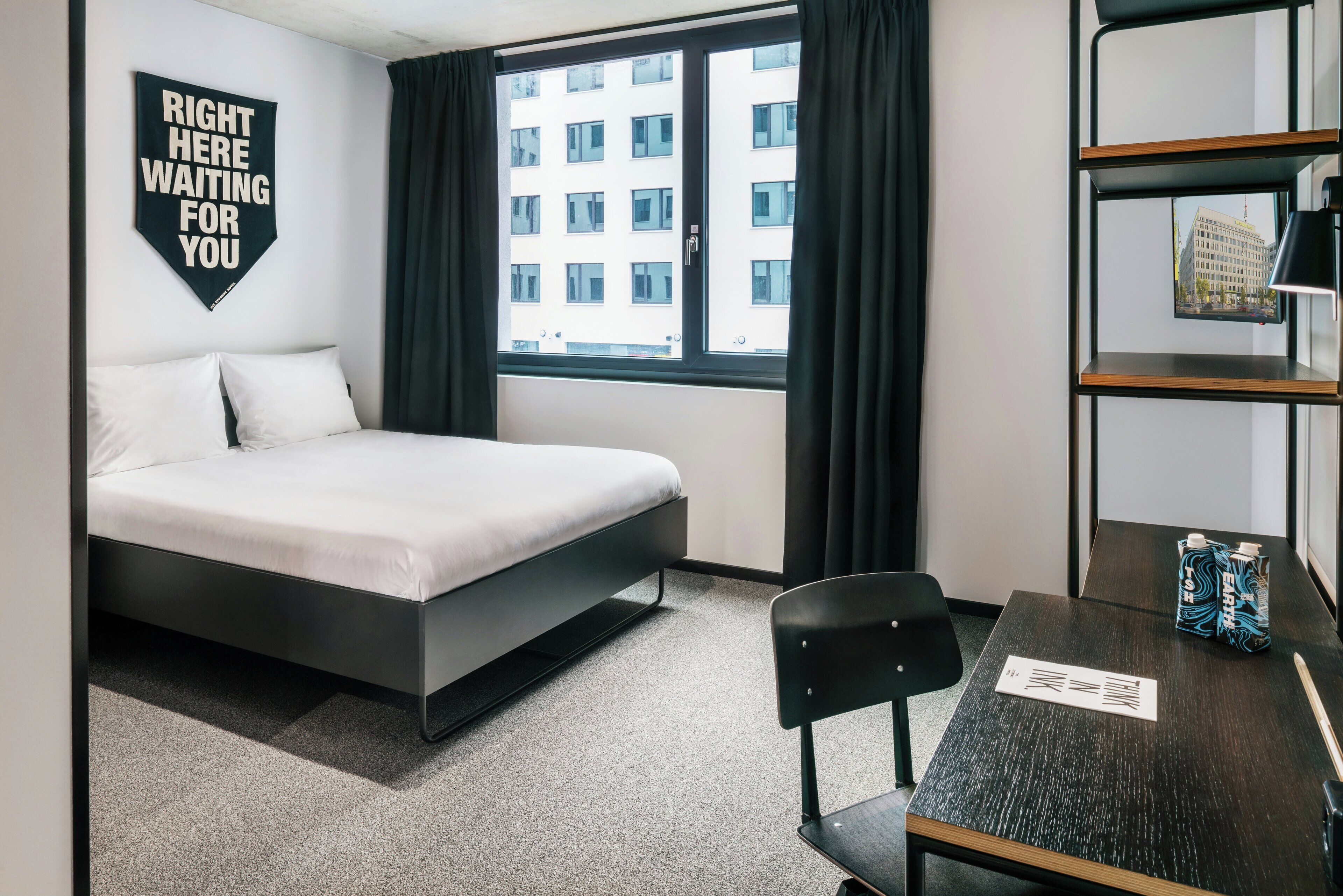 A modern hotel room with a bed, a desk, a chair, a bookshelf, and a window with curtains.