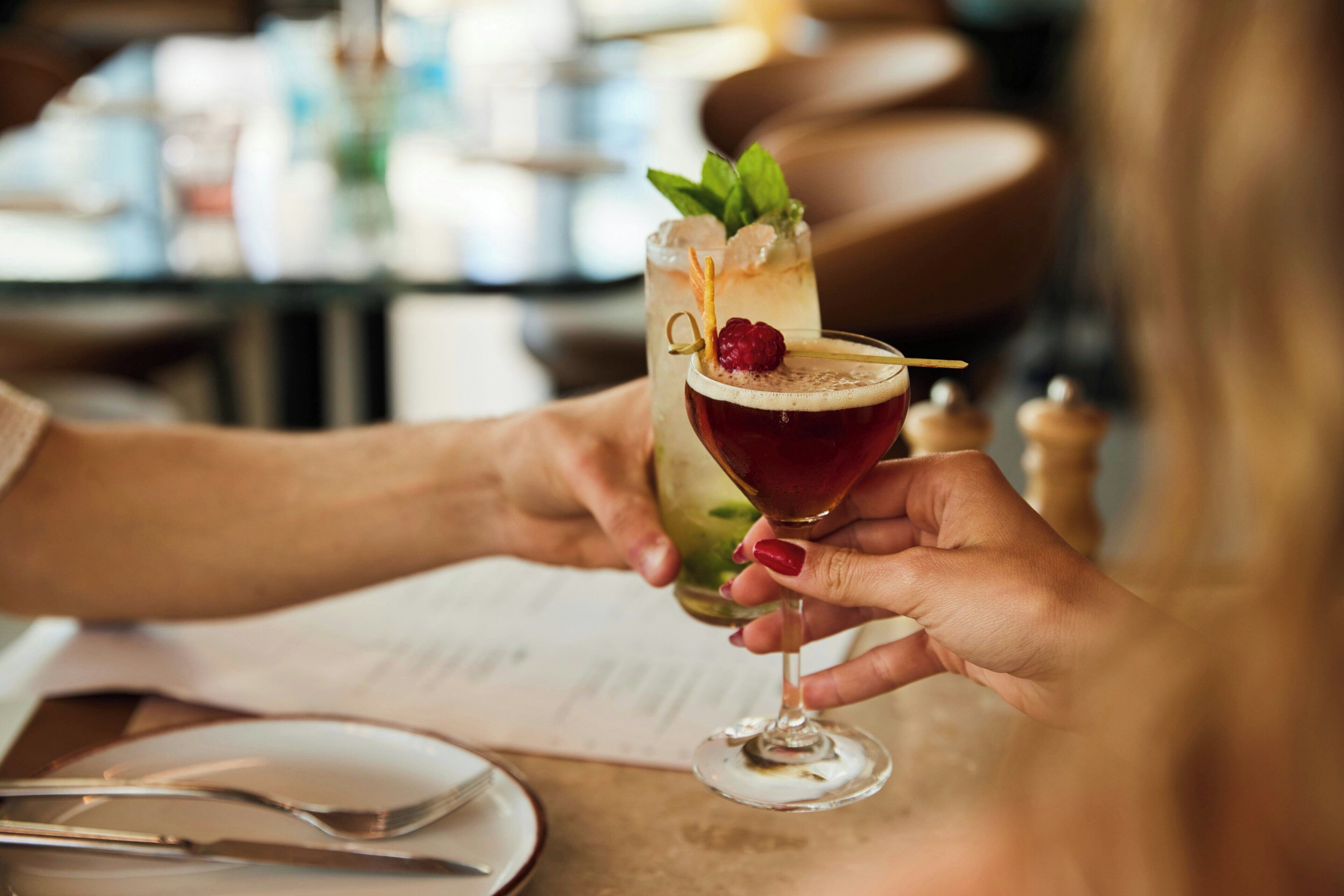 Zwei Hände halten Cocktailgläser mit Garnitur, eine Speisekarte und eine Gabel liegen auf dem Tisch.