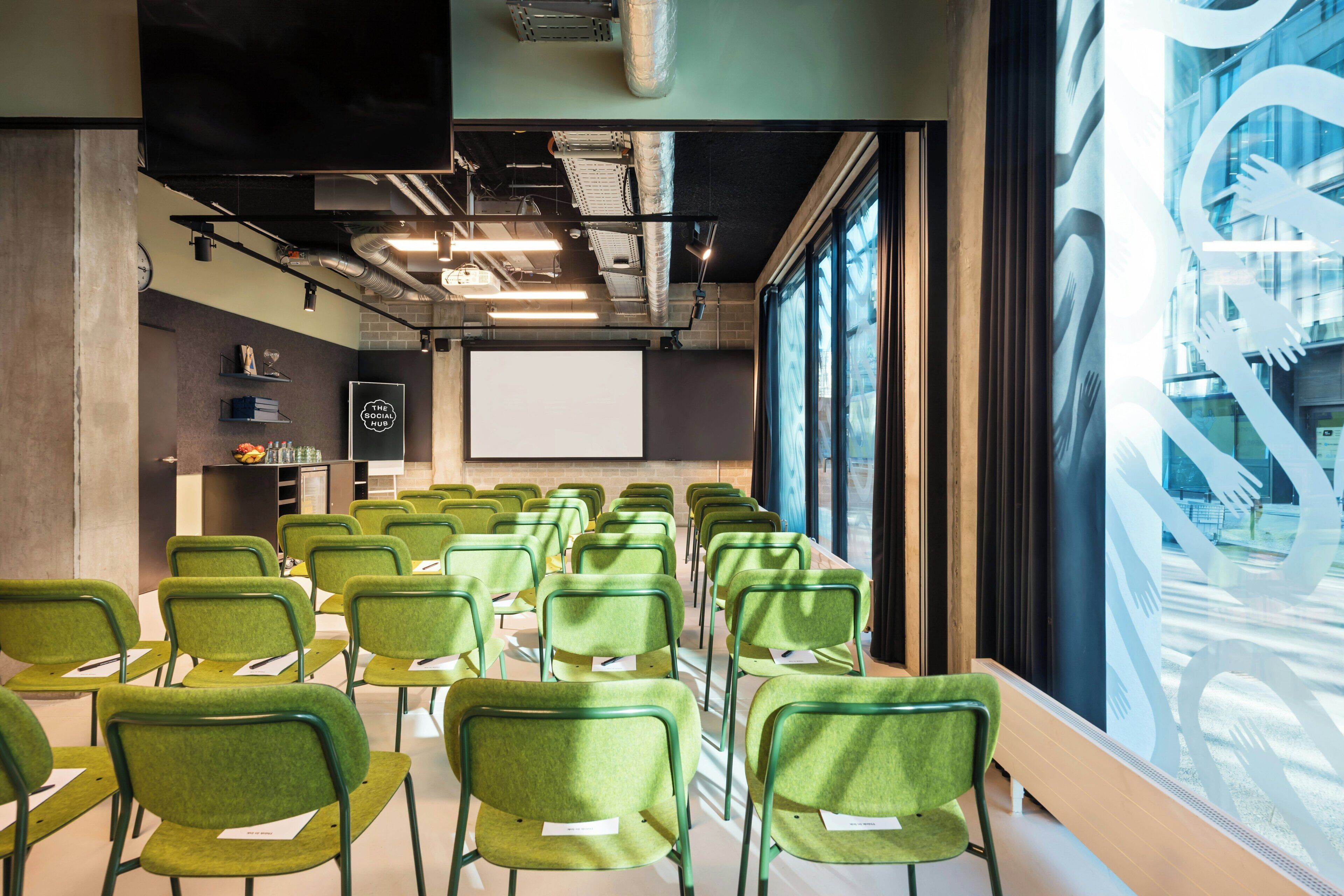 A conference room with rows of green chairs, a projector screen, and a large window with abstract art.