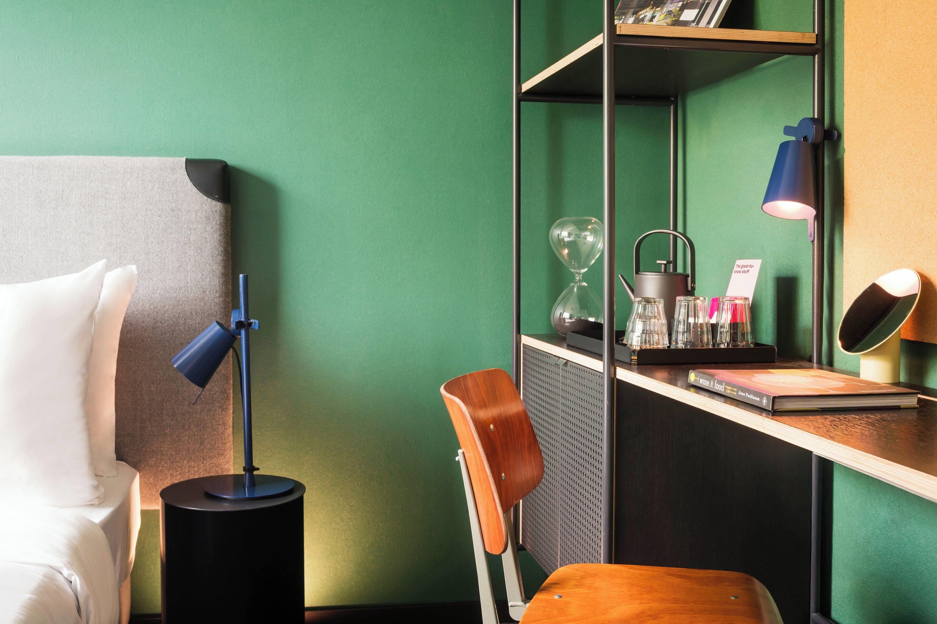 A hotel room with a bed, a desk with a lamp, a chair, and a shelf with various items.