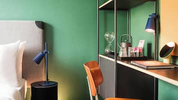 A hotel room with a bed, a desk with a lamp, a chair, and a shelf with various items.