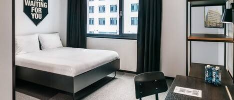 A modern hotel room with a bed, a desk, a chair, and a window with a view of a building.