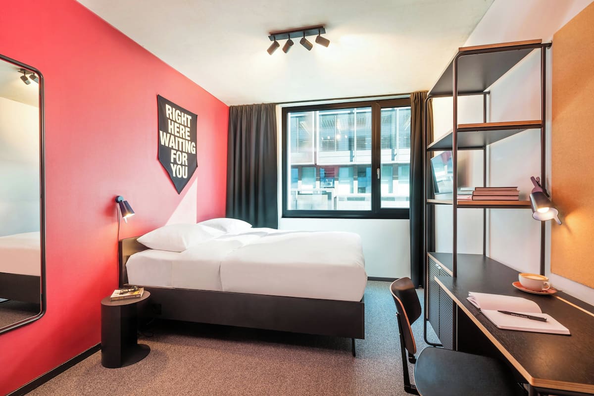 A hotel room with a bed, a desk, a chair, a mirror, and a window with curtains.