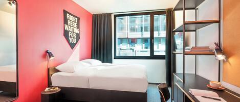 A hotel room with a bed, a desk, a chair, a mirror, and a window with curtains.